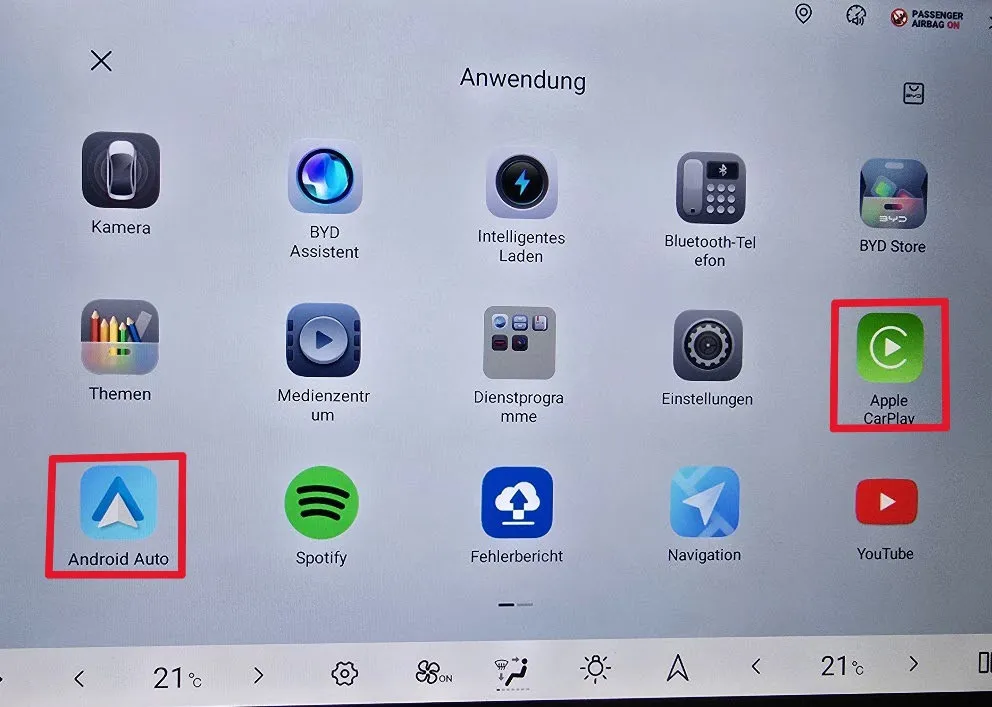 Head-unit app drawer titled Anwendung showing Kamera, BYD Assistent, Intelligentes Laden, Bluetooth-Telefon, BYD Store, Themen, Medienzentrum, Dienstprogramme, Einstellungen, Apple CarPlay, Android Auto, Spotify, Fehlerbericht, Navigation and YouTube, with Android Auto and Apple CarPlay each ringed in red.