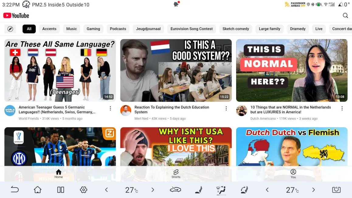YouTube running full screen on the head unit, showing the home feed with category chips (All, Accents, Music, Gaming, Podcasts) and video thumbnails, with the climate and shortcut bar along the bottom of the display.